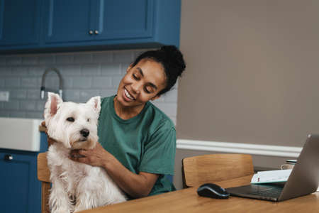 Smiling mid aged african woman sitting at the table indoors with her dog on a video call via laptop