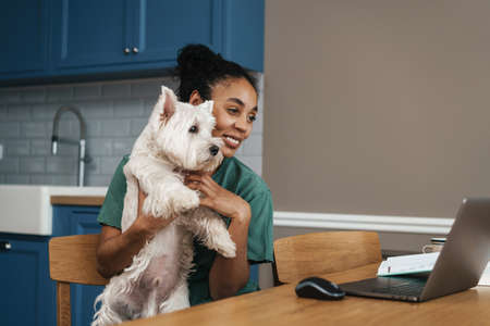 Smiling mid aged african woman sitting at the table indoors with her dog on a video call via laptopの写真素材