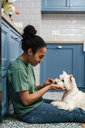 Smiling black woman feeding her dog while sitting on floor at home kitchenの写真素材