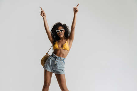 Black woman in swimsuit smiling and pointing fingers upward isolated over white backgroundの写真素材