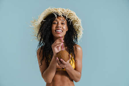 Black woman in straw hat smiling while drinking coconut water isolated over blue background