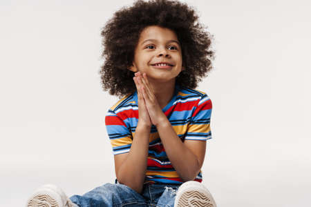 Happy preschooler african boy wearing t-shirt sitting over white wall, looking awayの写真素材