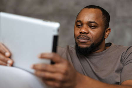 Black unshaven man using tablet computer while sitting on sofa at homeの写真素材
