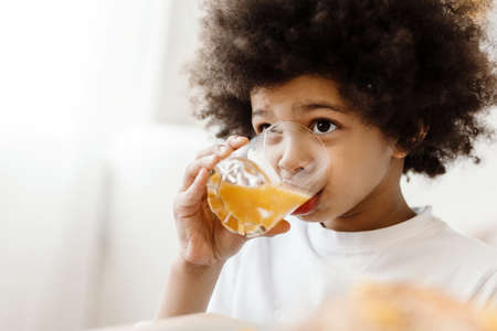 Black curly boy looking forward while drinking juice indoorsの写真素材