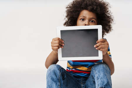 Preschooler african boy wearing t-shirt sitting over white wall, showing empty digital tablet...