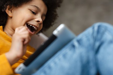 Black curly boy smiling and using tablet computer while sitting on sofa at homeの写真素材