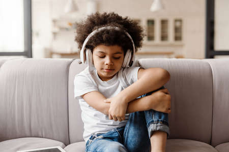Black curly boy listening music with headphones while sitting on sofa at homeの写真素材