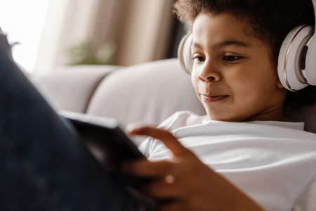 Black curly boy in headphones using tablet computer while sitting on sofa at homeの写真素材