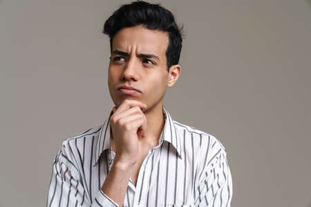 Brunette hispanic man in shirt frowning and looking aside isolated over grey background