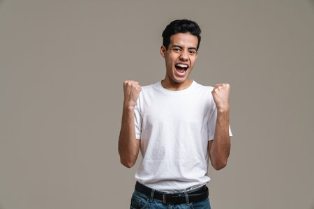 Smiling young brunette man in t-shirt standing over gray wall background, celebrating success