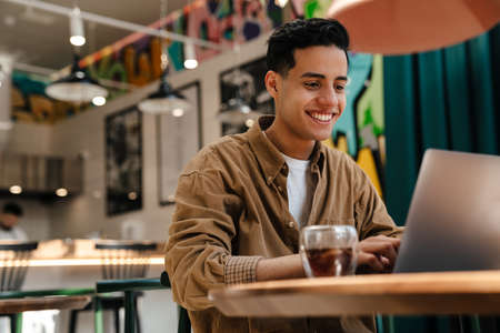 Young hispanic man student sitting at the cafe table with laptop computer indoors
