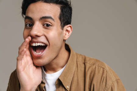Smiling excited young brunette man in t-shirt standing over gray wall background, looking at camera, holding hand at his open mouthの写真素材