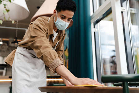 Young hispanic waiter disinfecting table in cafe while wearing face maskの写真素材