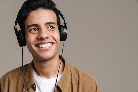 Smiling young brunette man in t-shirt standing over gray wall background, listening to music with headphonesの写真素材