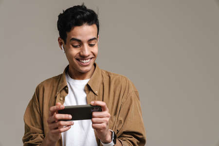 Smiling young brunette man in t-shirt standing over gray wall background, holding mobile phone,...