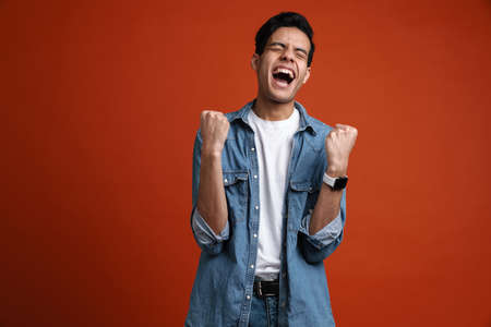 Excited hispanic man in shirt screaming while making winner gesture isolated over red backgroundの写真素材