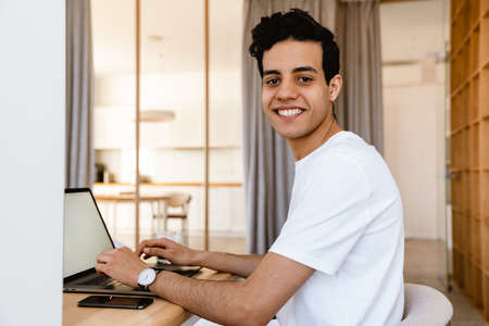 Smiling young hispanic man working studying on laptop computer while sitting at the desk at home