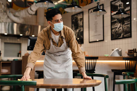Young hispanic waiter disinfecting table in cafe while wearing face maskの写真素材