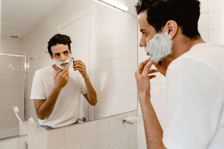 Young hispanic man shaving his face standing at the mirror indoorsの写真素材