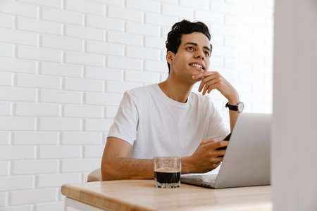 Smiling young hispanic man working studying on laptop computer while sitting at the desk at homeの写真素材