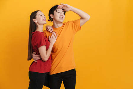Young multiracial couple smiling and hugging while looking upward isolated over yellow wall