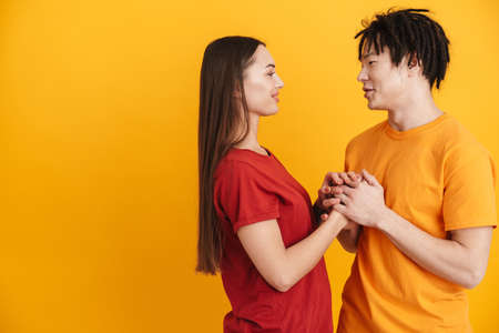 Young multiracial couple smiling and holding hands together while talking isolated over yellow wall