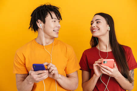 Young multiracial couple listening music with earphones and cellphones isolated over yellow wallの写真素材