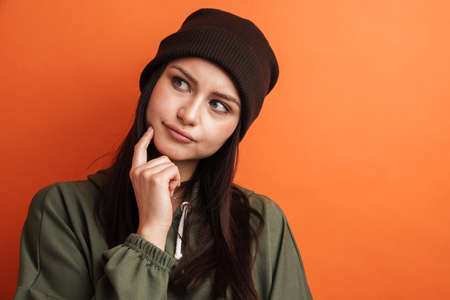 Pensive young casual brunette woman with long hair wearing hat standing over red background, looking asideの写真素材