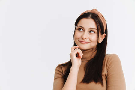 Happy pensive young brunette white woman in casual clothes standing over white background, looking...