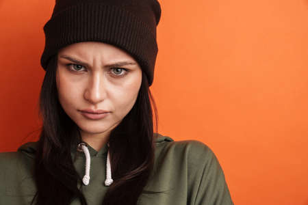 Pensive frowning young casual brunette woman with long hair wearing hat standing over red background, looking at cameraの写真素材