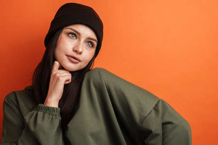 Pensive young casual brunette woman with long hair wearing hat standing over red background, looking asideの写真素材