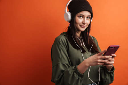 Happy young casual brunette woman with long hair wearing hat standing over red background, wearing headphones, listening to music, holding mobile phoneの写真素材