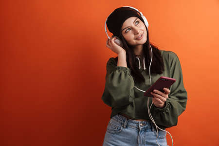 Happy young casual brunette woman with long hair wearing hat standing over red background, wearing...