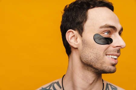 Close up of a shirtless mid aged brunette man wearing cosmetic eye patches over yellow wall backgroundの写真素材