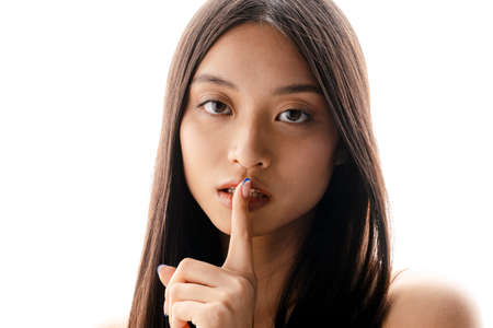 Young asian brunette woman looking and showing silence gesture at camera isolated over white wallの写真素材
