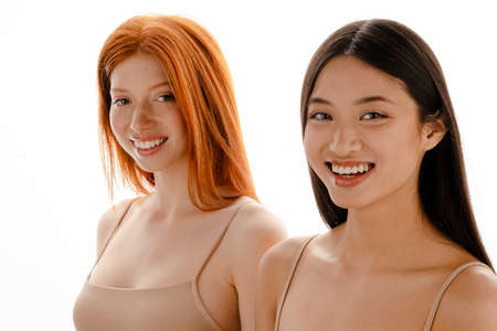 Multiracial two women looking and laughing at camera isolated over white wall