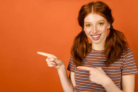 Ginger cheerful woman with tails smiling and pointing fingers aside isolated over orange backgroundの写真素材