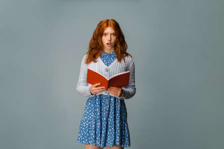 Young ginger shocked woman posing with exercise book isolated over grey background