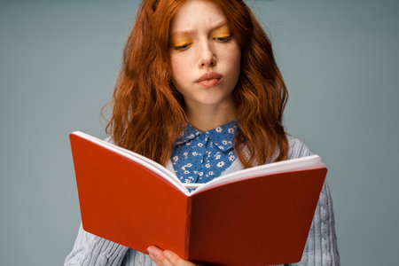 Young ginger puzzled woman posing while reading her diary isolated over grey background