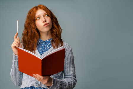 Young ginger thinking woman posing with exercise book and pencil isolated over grey backgroundの写真素材