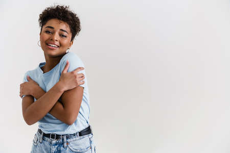 Smiling excited young african woman in casual wear standing over white wall, looking at camera, hugging herselfの写真素材