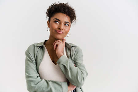 Smiling pensive young african woman in casual wear standing over white wall