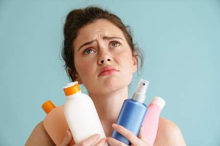 Half-naked brunette woman frowning while posing with beauty products isolated over blue backgroundの写真素材