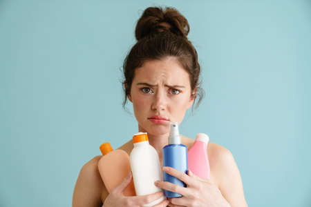 Half-naked brunette woman frowning while posing with beauty products isolated over blue backgroundの写真素材