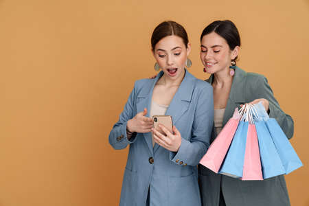 Young two women holding shopping bags and using cellphone isolated over yellow backgroundの写真素材