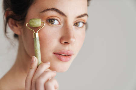 brunette woman massaging face with jade roller isolated over while backgroundの写真素材