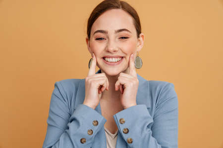 Young ginger woman smiling and pointing fingers at her cheeks isolated over yellow backgroundの写真素材