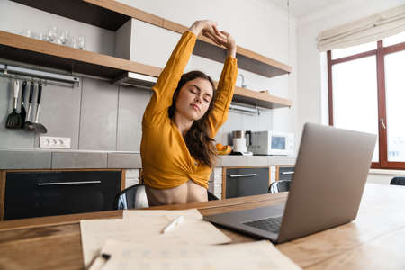 Smiling young brunette woman working on laptop computer while sitting in the kitchen, stretching...