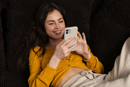 Smiling young brunette woman using mobile phone while laying on a couchの写真素材