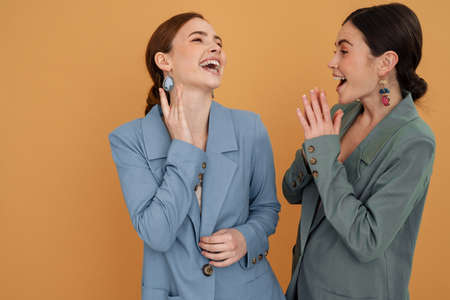 Two smiling elegant women wearing smart jackets posing standing close together isolated over beige background laughingの写真素材